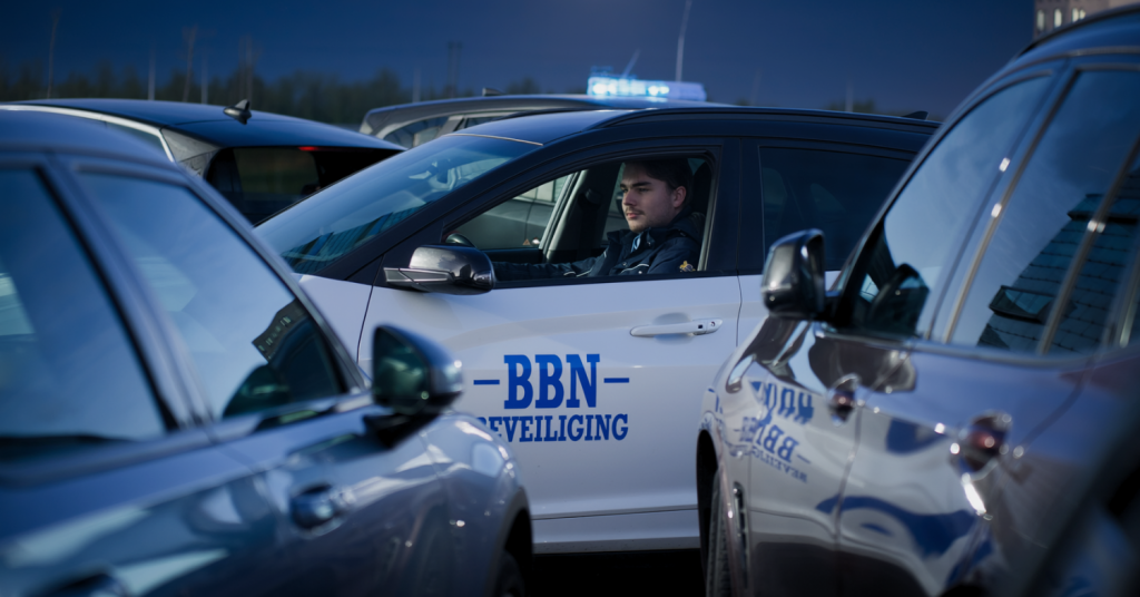 Beveiliger van BBN Beveiliging voert in de avonduren surveillance uit op een parkeerterrein tussen geparkeerde voertuigen. De surveillant draagt bedrijfskleding met zichtbaar BBN-logo en houdt toezicht ter ondersteuning van veiligheid, preventie en collectieve surveillance.